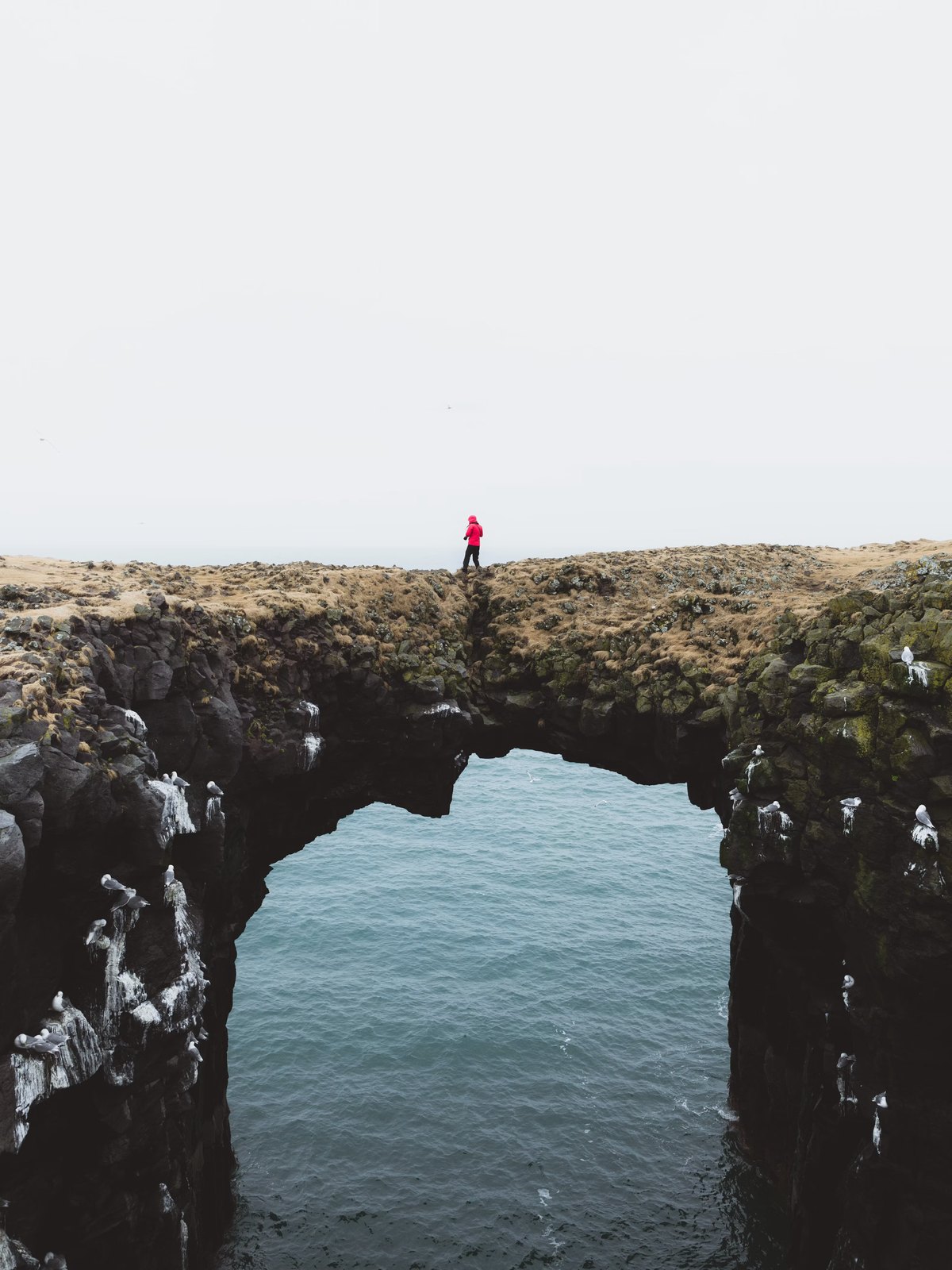 A lone figure standing on a natural rock arch above the sea
